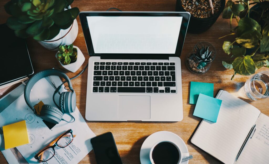 Overhead view of a laptop, headphones, plants, notebook, and coffee on a wooden desk.