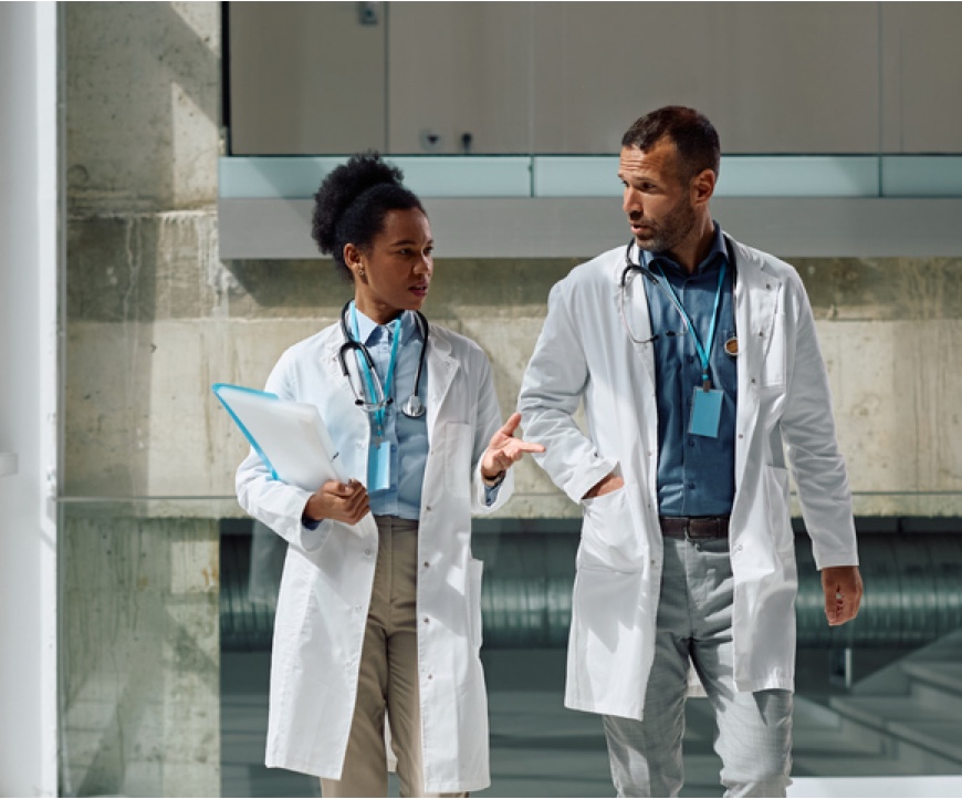 Two doctors in lab coats walk and talk, one holding a clipboard, in a modern hallway.