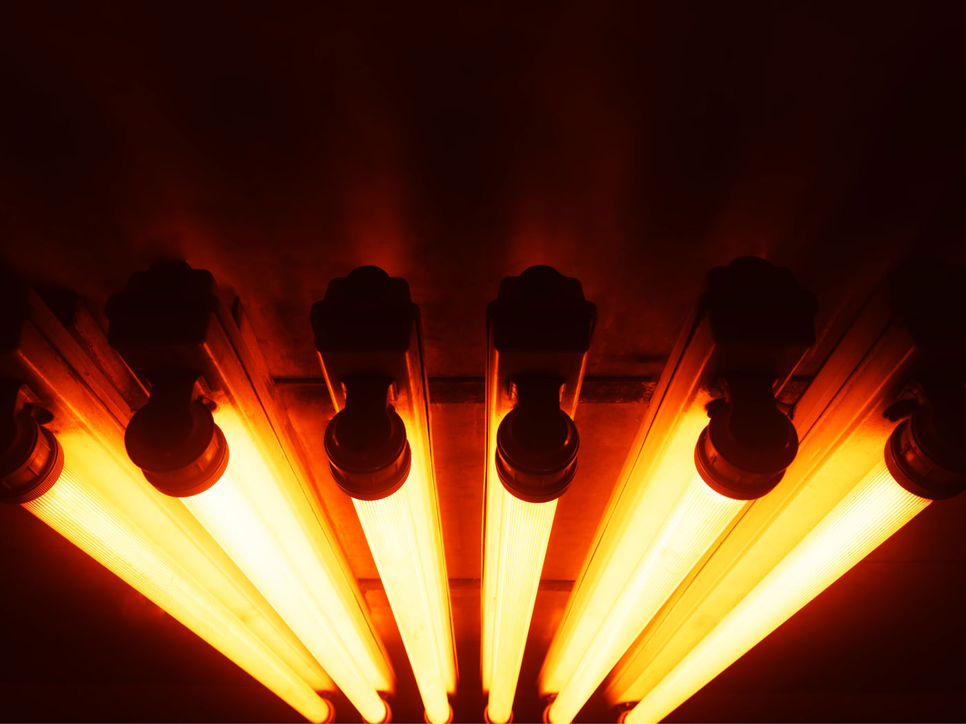 Four orange fluorescent light tubes glowing on a dark ceiling, casting warm amber illumination.