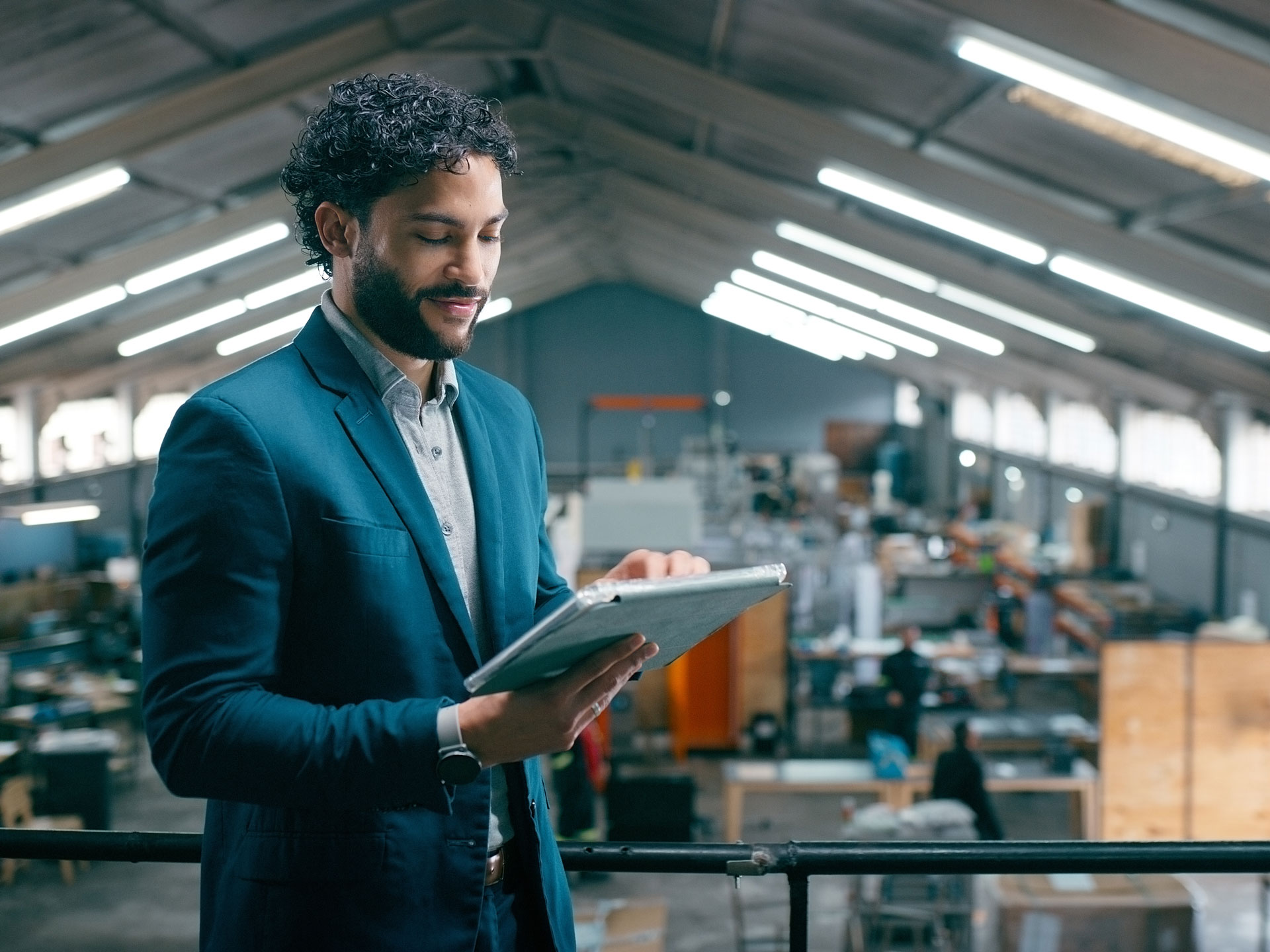 Curly-haired man in teal suit holding a tablet on a balcony overlooking a busy industrial warehouse with fluorescent lights
