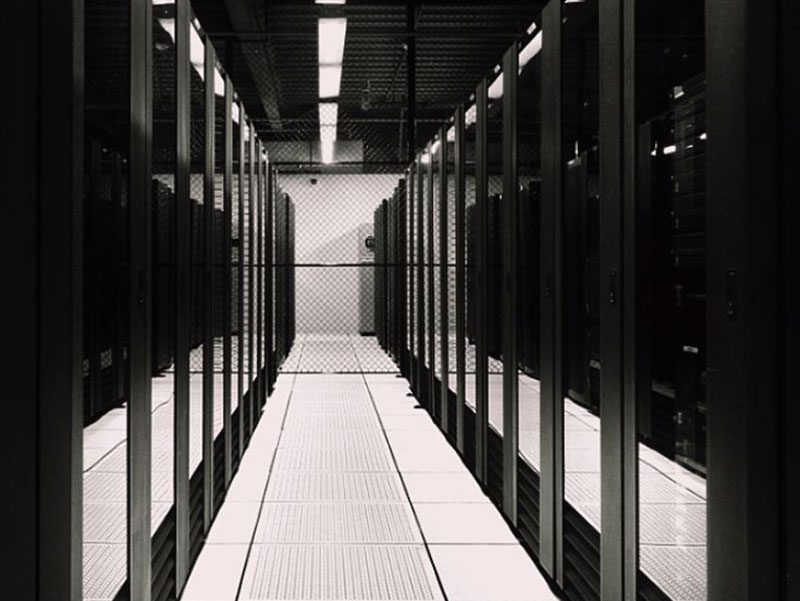 Black and white view down aisle in server room with rows of server racks and chain-link fence at end.