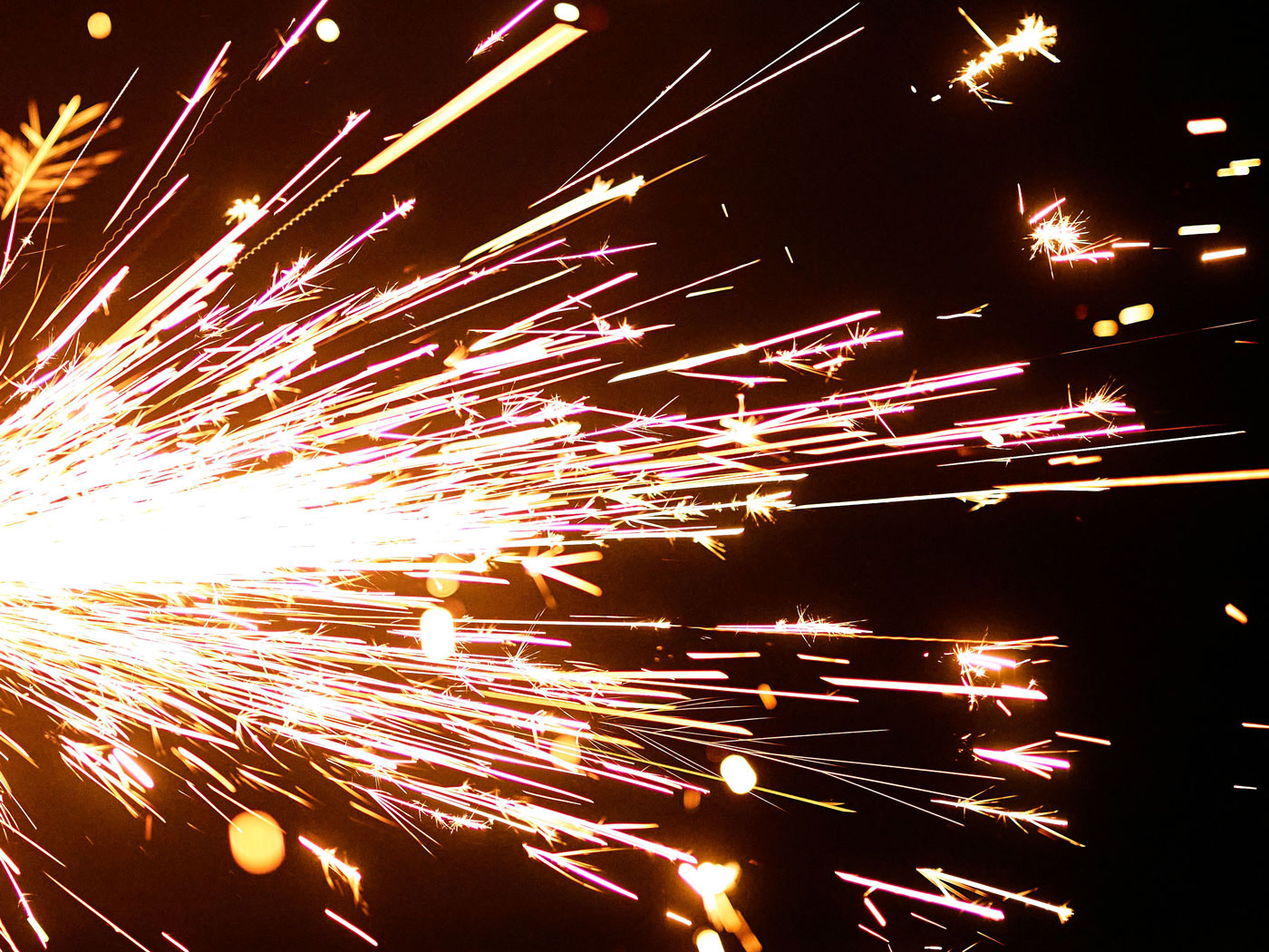 Bright golden and pink sparks exploding from a firework against a black night sky, creating dynamic light trails.