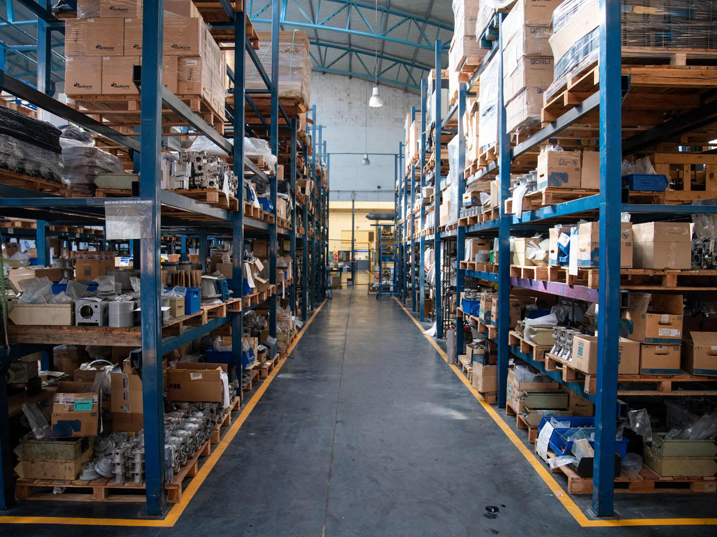 Wide aisle in a warehouse with blue metal racks filled with boxes, pallets and industrial parts on a concrete floor.