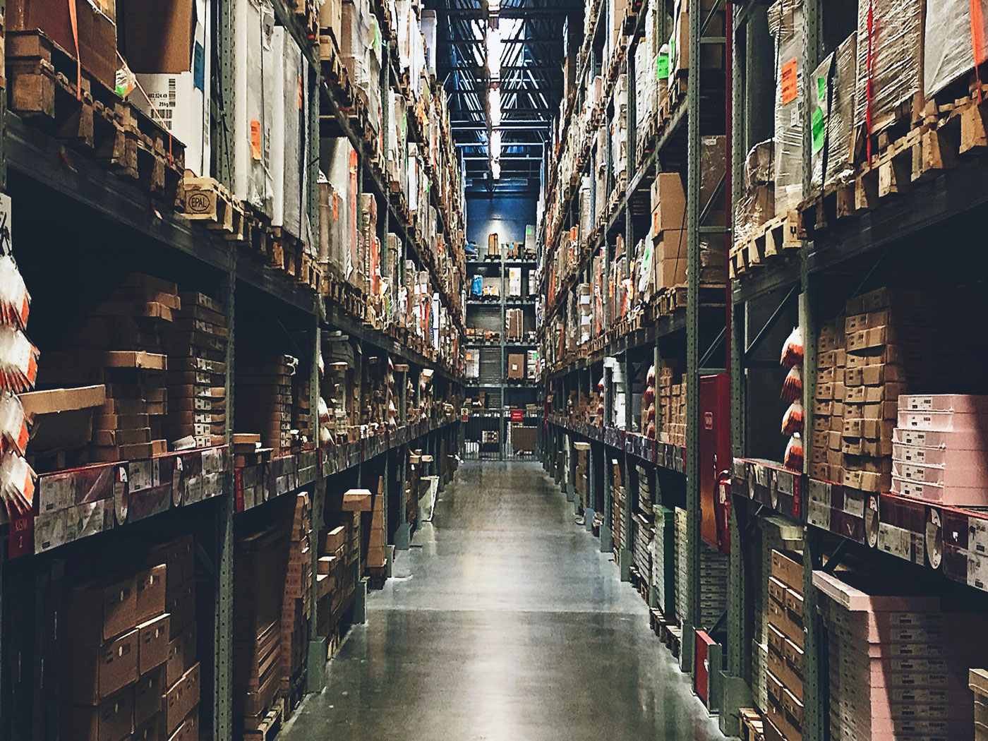 Long warehouse aisle with tall metal shelves stacked with cardboard boxes and pallets under bright lights.