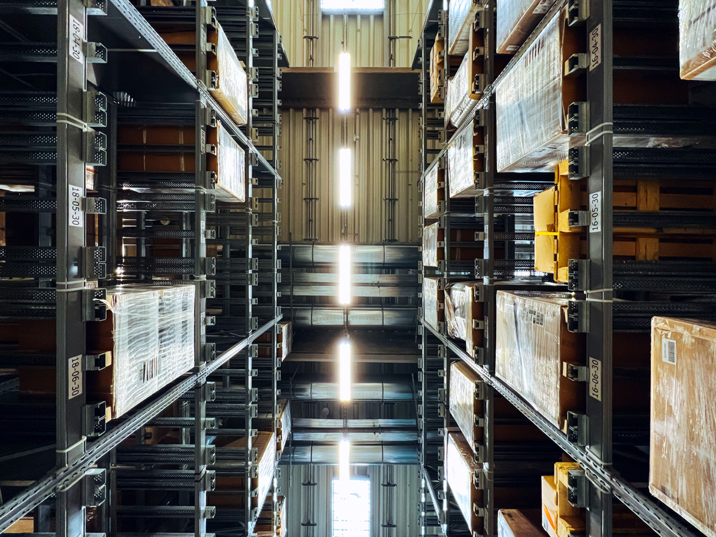 Vertical view of tall warehouse metal shelves loaded with boxes and pallets, lit by fluorescent lights.