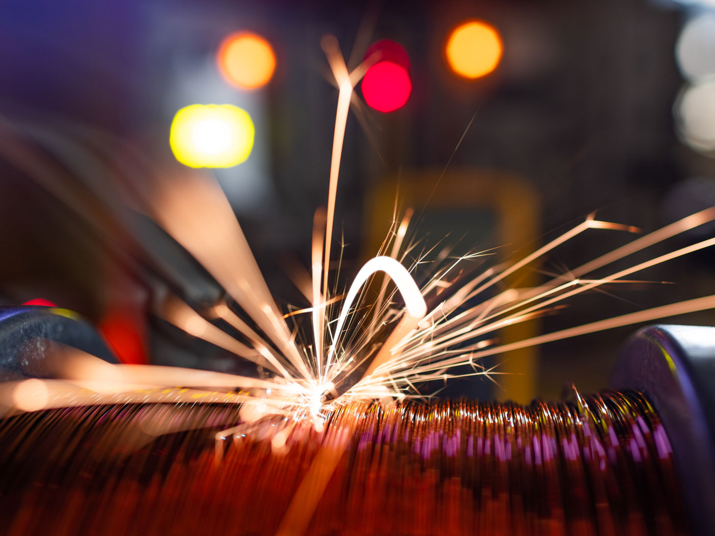 Bright sparks fly from a welding torch as it fuses copper wire, with colorful blurred lights in the background.