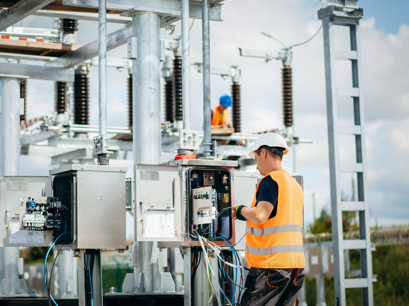 Two electricians in hard hats and safety vests work on electrical panels in a power substation, handling wires and equipment.