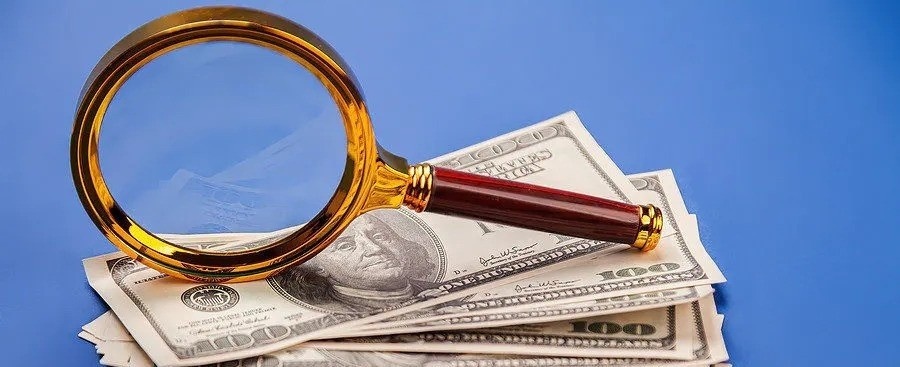 Gold-rimmed magnifying glass inspecting a stack of US $100 bills on blue background